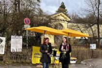 Zu sehen ist ein Farbfoto. Auf diesem werden abgebildet: Ein knallgelber Stand von Amnesty International auf einem Gehweg in Münchne. Vor dem Stand stehen zwei weiblich gelesene Personen. Beide Personen lächeln, halten weiße Rosen in den Händen.