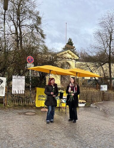 Zu sehen ist ein Farbfoto. Auf diesem werden abgebildet: Ein knallgelber Stand von Amnesty International auf einem Gehweg in Münchne. Vor dem Stand stehen zwei weiblich gelesene Personen. Beide Personen lächeln, halten weiße Rosen in den Händen.